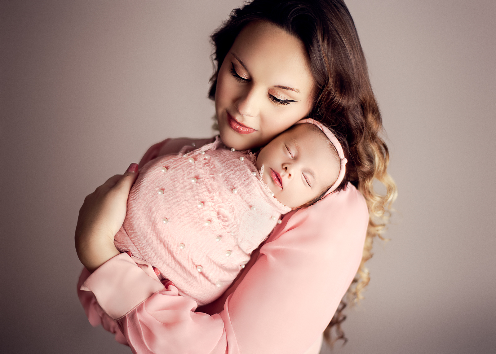 elegant mother and baby daughter dressed in soft pink, with baby bundled in a swaddled overlaid with pearls, cuddled on mom's shoulder